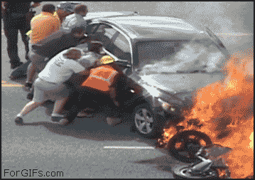 A group of people help lift a burning car to pull out a trapped man from under it