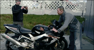 A guy revs a motorcycle while standing in front of it and it drives him into a shed behind him